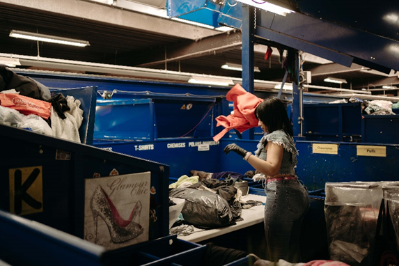 1. Une femme trie des vêtements dans un centre de tri textile, entourée de piles de vêtements colorés.